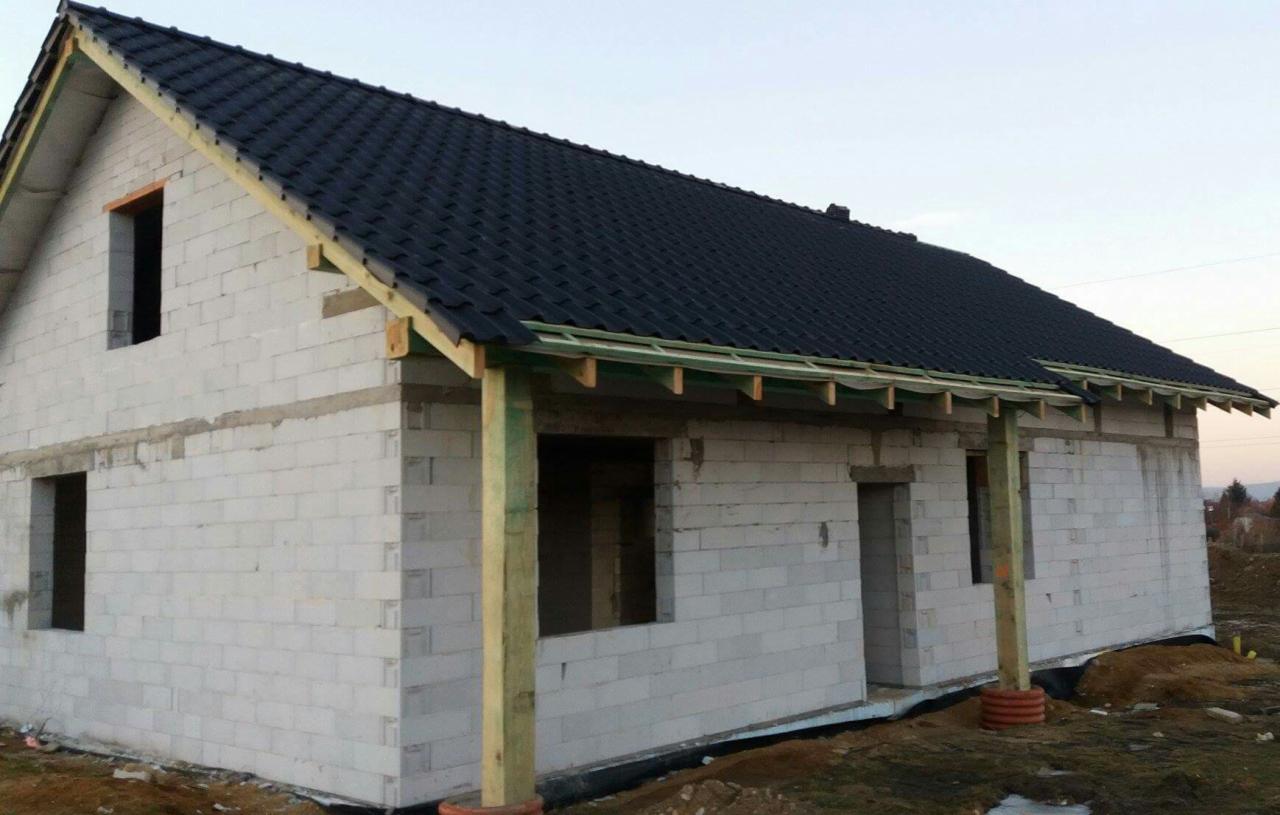 Surowy stan domu jednorodzinnego z białych bloczków, z ciemną dachówką i drewnianą konstrukcją zadaszenia werandy.