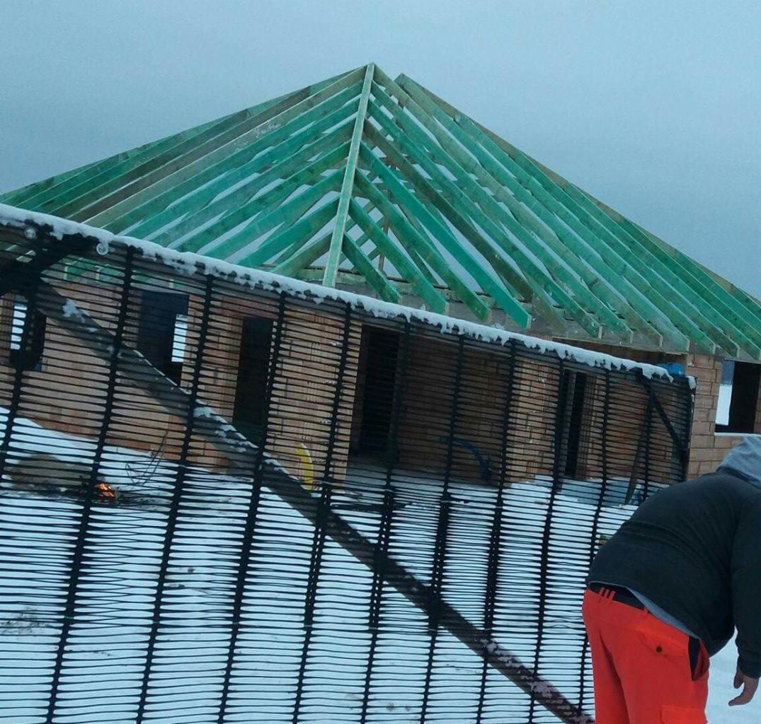 Wznoszona konstrukcja domu murowanego z zielonymi elementami więźby dachowej, widoczna przez siatkę ochronną, z fragmentem sylwetki robotnika w pomarańczowych spodniach w prawym dolnym rogu.