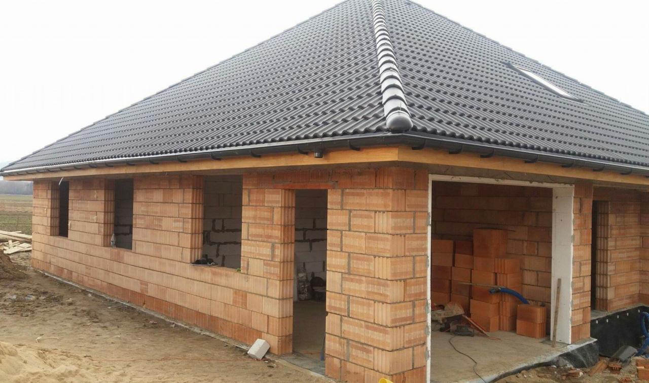 Surowy stan domu z cegły ceramicznej z ciemną dachówką, widoczne okna bez szyb i drzwi, wewnątrz składowane cegły.