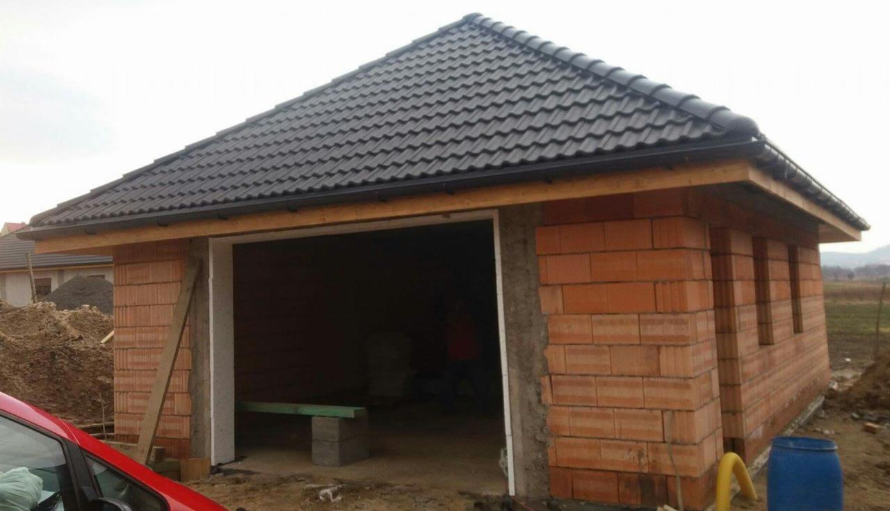 Surowy budynek garażu z czerwonej cegły ceramicznej z czarnym dachem z dachówki, widok z zewnątrz na etapie budowy, w tle widoczne wzgórza i fragment czerwonego samochodu.