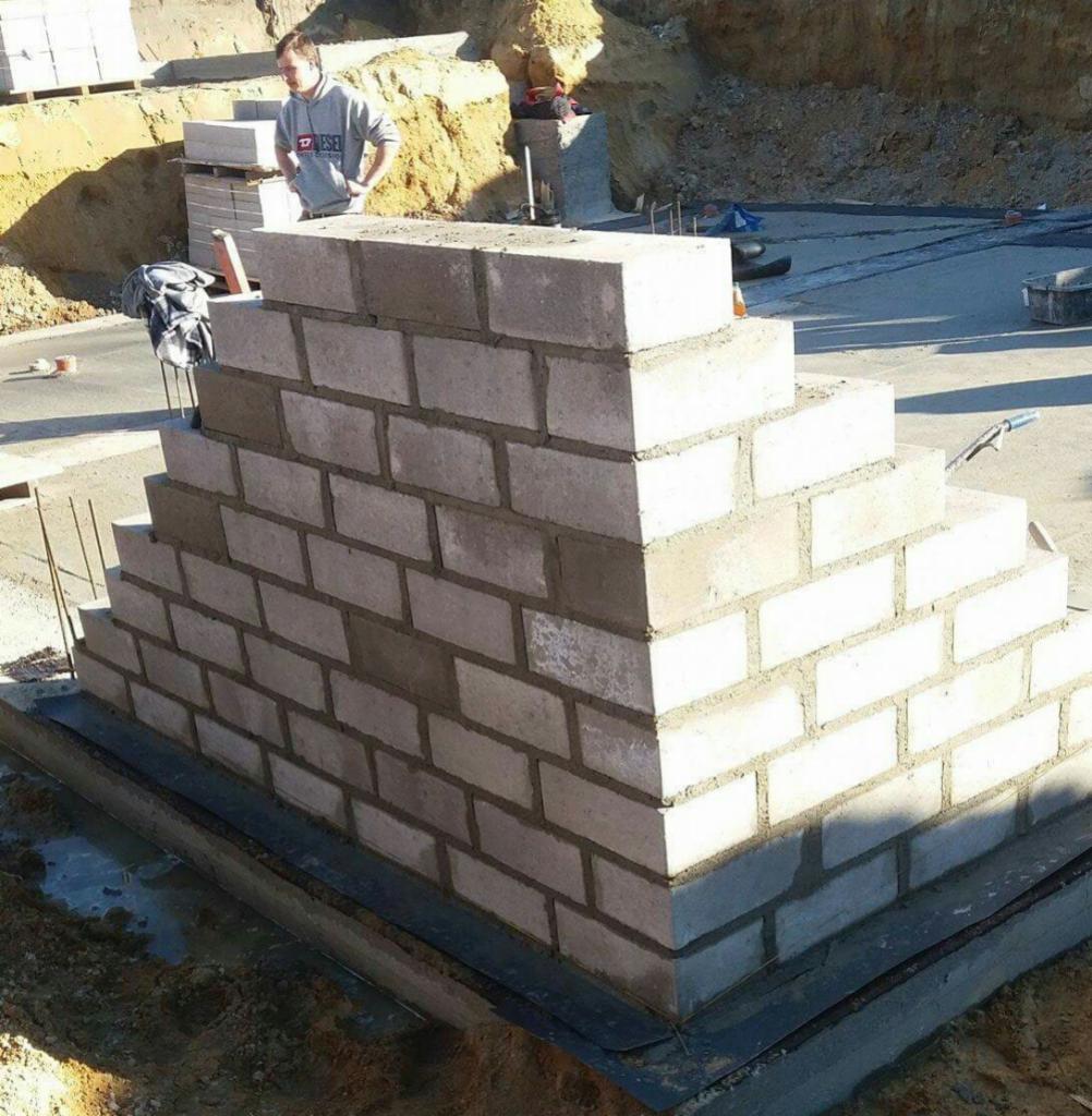 Wznoszenie piramidalnej konstrukcji z białych bloczków betonowych na fundamencie, widoczny murarz w tle i przygotowane materiały budowlane