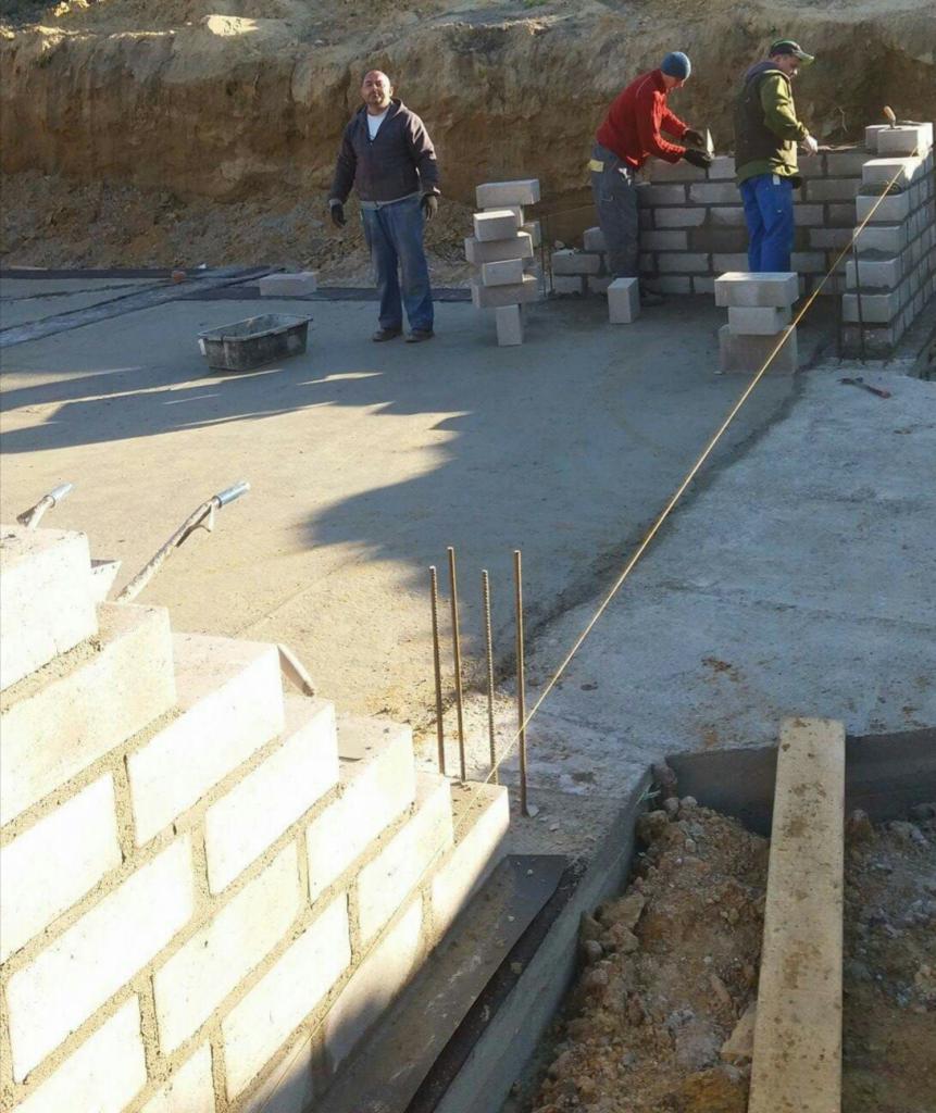 Budowa ściany z cegły na betonowym fundamencie; trzech pracowników w roboczych ubraniach, jeden obserwuje, dwóch muruje cegły; widoczne stalowe pręty zbrojeniowe wystające z fundamentu, sznurek...