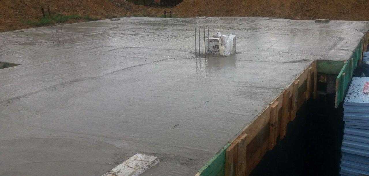 Świeżo wylana, mokra płyta fundamentowa z wystającymi prętami zbrojeniowymi, otoczona drewnianym szalunkiem i izolacją ze styropianu.