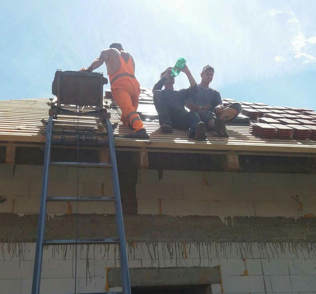 Przerwa na dachu: Trzech robotników podczas układania dachówki, jeden w pomarańczowym kombinezonie przy windzie dekarskiej, dwaj odpoczywają i piją wodę z butelki, widoczna surowa ściana budynku...