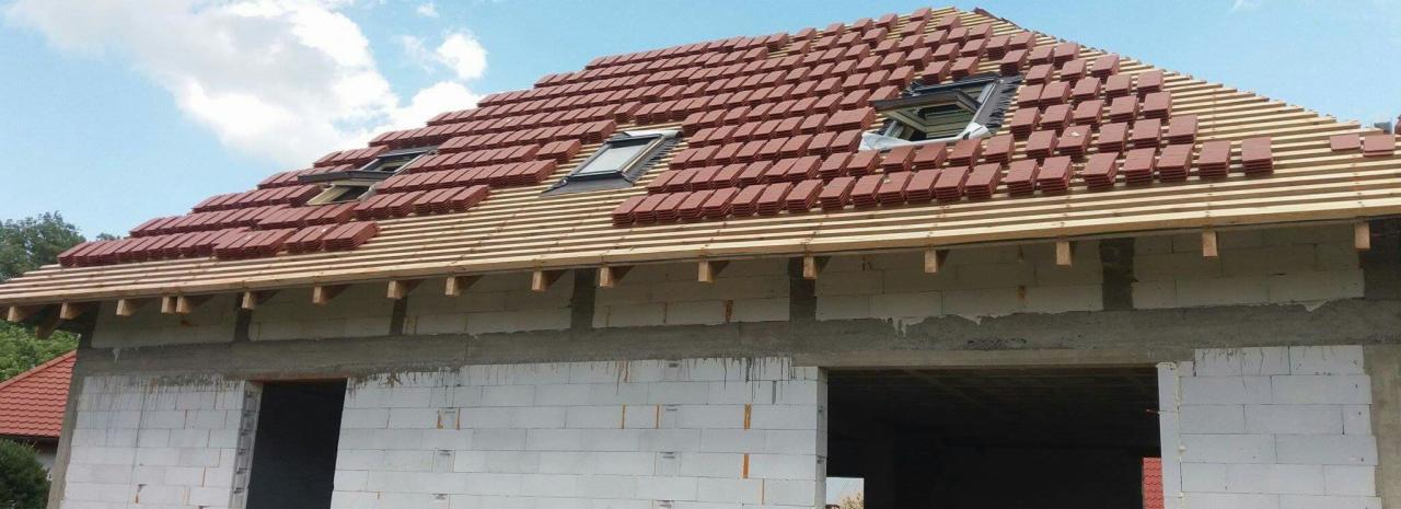 Wznoszenie dachu z dachówką ceramiczną na surowym budynku z białego bloczka, widoczne okna dachowe w trakcie montażu.