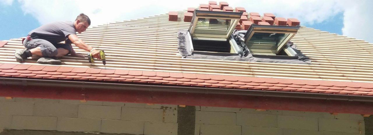 Montaż dachówki karpiówki na drewnianej konstrukcji dachu z oknami dachowymi, wykonywany przez dekarza z użyciem wkrętarki.