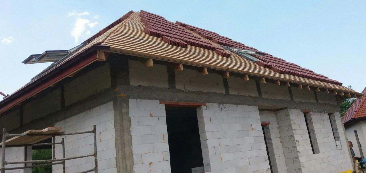 Budowa domu z białych bloczków: widoczne ściany w trakcie wznoszenia, częściowo ułożona dachówka w kolorze czerwonym, okno dachowe, drewniana konstrukcja dachu.