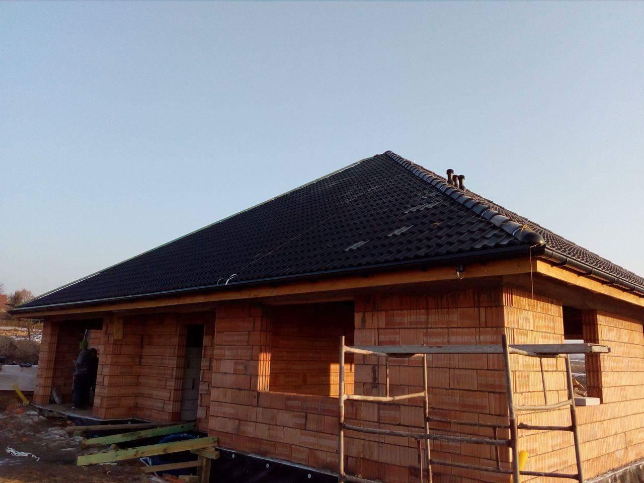 Surowy stan domu jednorodzinnego z czerwonej cegły z czarnym dachem, widoczne rusztowanie i brak okien, w tle błękitne niebo.