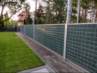 Nowoczesne ogrodzenie panelowe z siatką maskującą w kolorze zielonym, oddzielające posesję z trawnikiem od sąsiedniej działki z domem. Widoczne drzewa w tle.