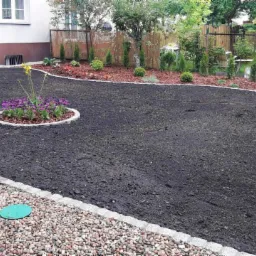 Przygotowany teren pod trawnik z obrzeżami z kostki, rabata z kwiatami na środku, w tle nasadzenia ozdobne z kory.