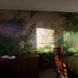 Wnętrze pokoju z tapetą przedstawiającą tropikalny krajobraz z oknem, drewnianą komodą i krzesłami w tle, oświetlone popołudniowym słońcem.