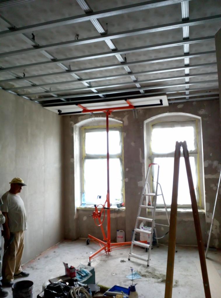 Montaż sufitu podwieszanego z wykorzystaniem podnośnika, widoczne profile i płyta gipsowo-kartonowa, pracownik w żółtej czapce i drabina w pomieszczeniu z dwoma oknami.