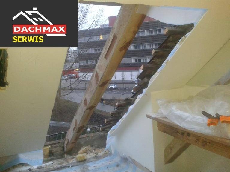 Uszkodzona konstrukcja okna dachowego z widoczną belką drewnianą i fragmentem budynku w tle, częściowo zabezpieczona folią, widoczne narzędzia.