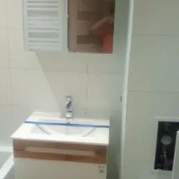 Nowoczesna łazienka z białą umywalką na szafce z drewnianymi akcentami, lustro z szafką wiszącą, grzejnik drabinkowy i płytki ceramiczne na ścianach, umywalka zabezpieczona taśmą malarską.