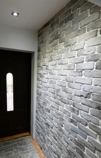 Ściana wewnętrzna wykończona szarą cegłą dekoracyjną, widok z perspektywy, obok ciemne drzwi z szybką w kształcie łuku.