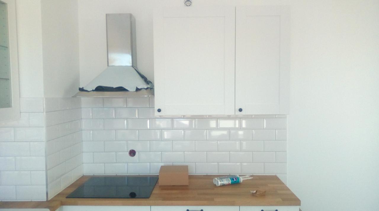 Fragment kuchni w trakcie wykańczania: białe, błyszczące płytki ułożone w cegiełkę, drewniany blat, częściowo zamontowane szafki wiszące i okap z folią ochronną, widoczne narzędzia i materiały.