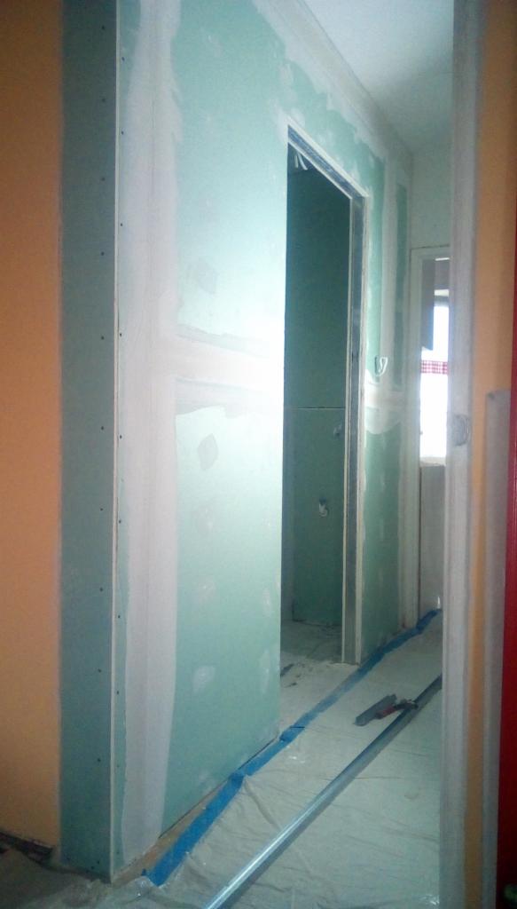 Wnętrze w trakcie prac wykończeniowych: ściany z płyt gipsowo-kartonowych, częściowo pokryte białą masą szpachlową, zabezpieczona folią podłoga z niebieską taśmą, widoczne narzędzia szpachlarskie.