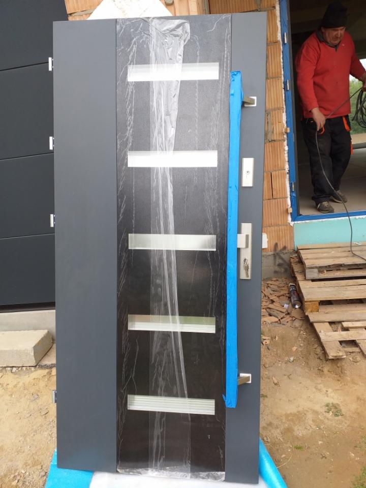 Szare drzwi zewnętrzne z przeszkleniami, zabezpieczone folią i taśmą malarską podczas montażu, widoczny pracownik w tle.