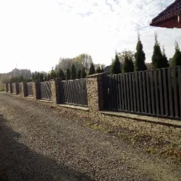 Nowoczesne ogrodzenie posesji z czarnymi, pionowymi sztachetami i kamiennymi słupkami oraz podmurówką, widok z drogi żwirowej.