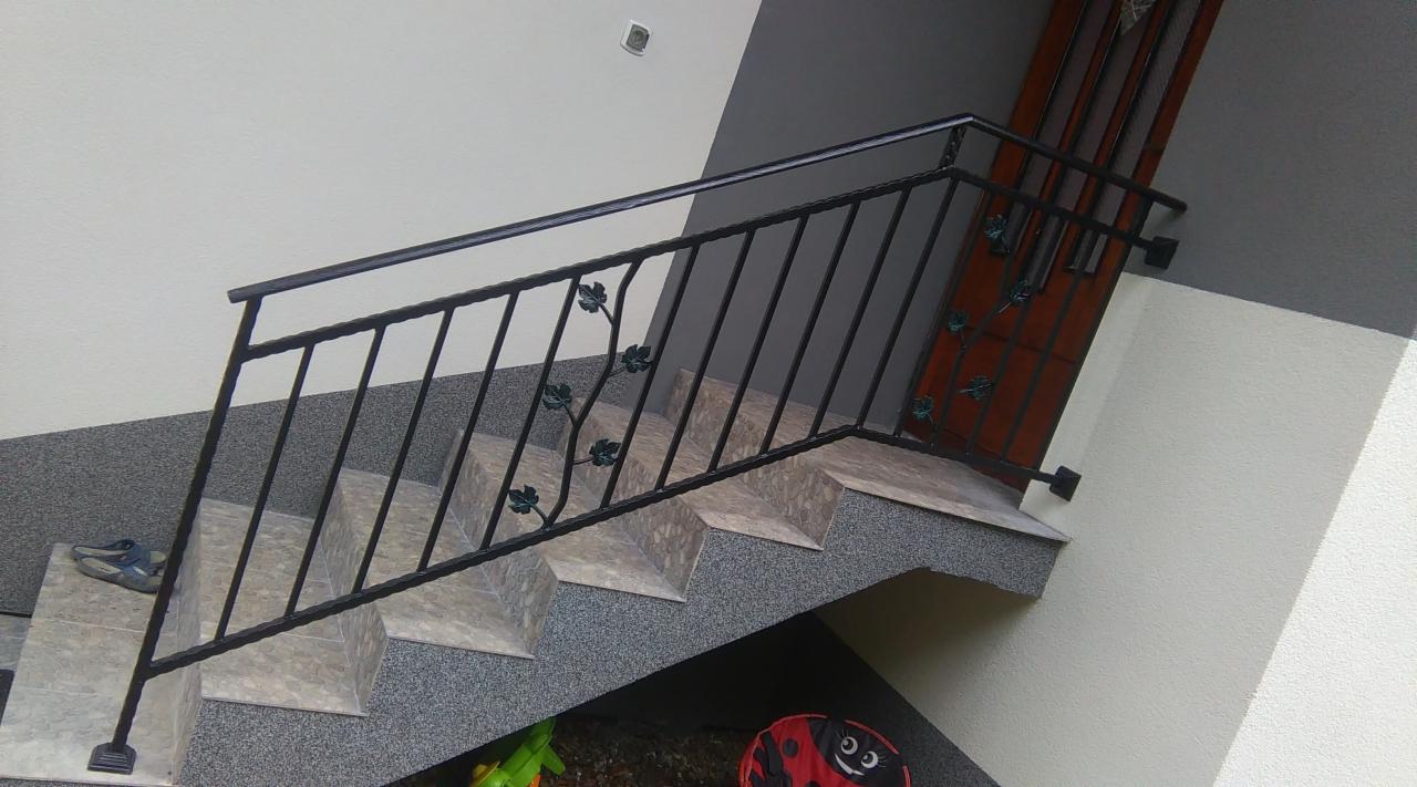 Klatka schodowa z czarną, kutą balustradą zdobioną motywem liści, prowadząca do drewnianych drzwi; na schodach leżą dziecięce buty i zabawki.