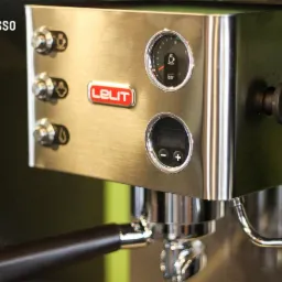 Szczegółowe ujęcie srebrnego ekspresu do kawy marki Lelit z manometrem, wyświetlaczem i dyszą pary na tle zielonej ściany.