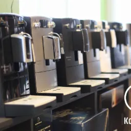 Rząd ekspresów do kawy w różnych odcieniach szarości i czerni, ustawionych na ciemnej półce w sklepie, z widocznym logo firmy Konesso na pierwszym planie.