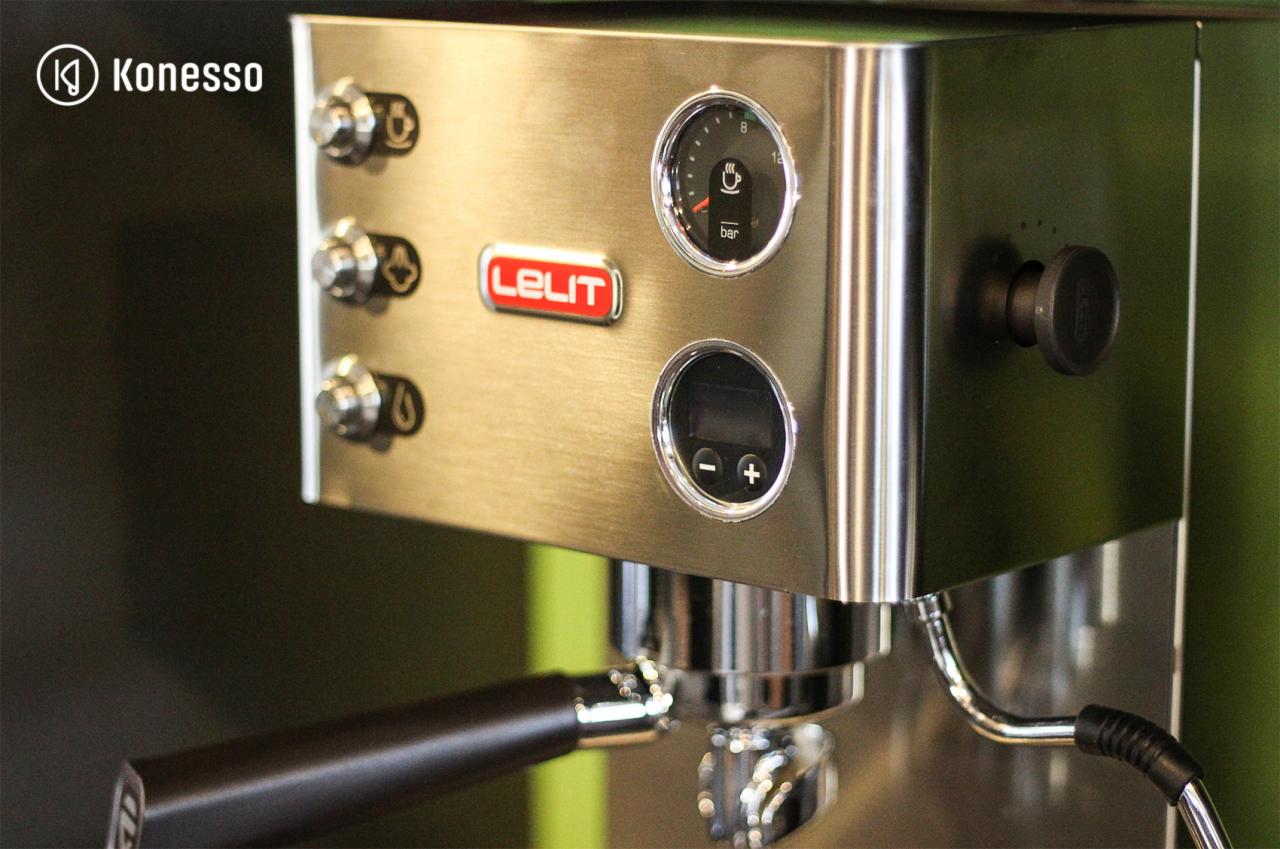 Szczegółowe ujęcie srebrnego ekspresu do kawy marki Lelit z manometrem, wyświetlaczem i dyszą pary na tle zielonej ściany.