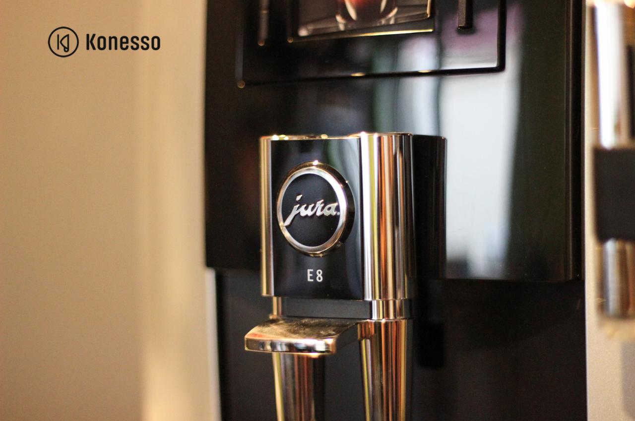Detale automatycznego ekspresu do kawy Jura E8 z chromowanymi elementami i logo Jura. Widoczny dyspenser kawy z regulowaną wysokością.