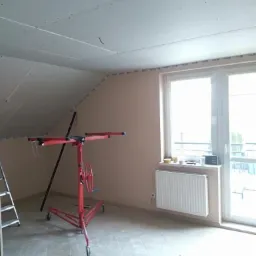 Montaż płyt na poddaszu z użyciem podnośnika, widoczne szpachlowane łączenia, okno z grzejnikiem w tle.