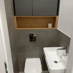 Nowoczesna toaleta z szarymi płytkami, wiszącą szafką z drewnianą wnęką i białą umywalką z chromowaną baterią.