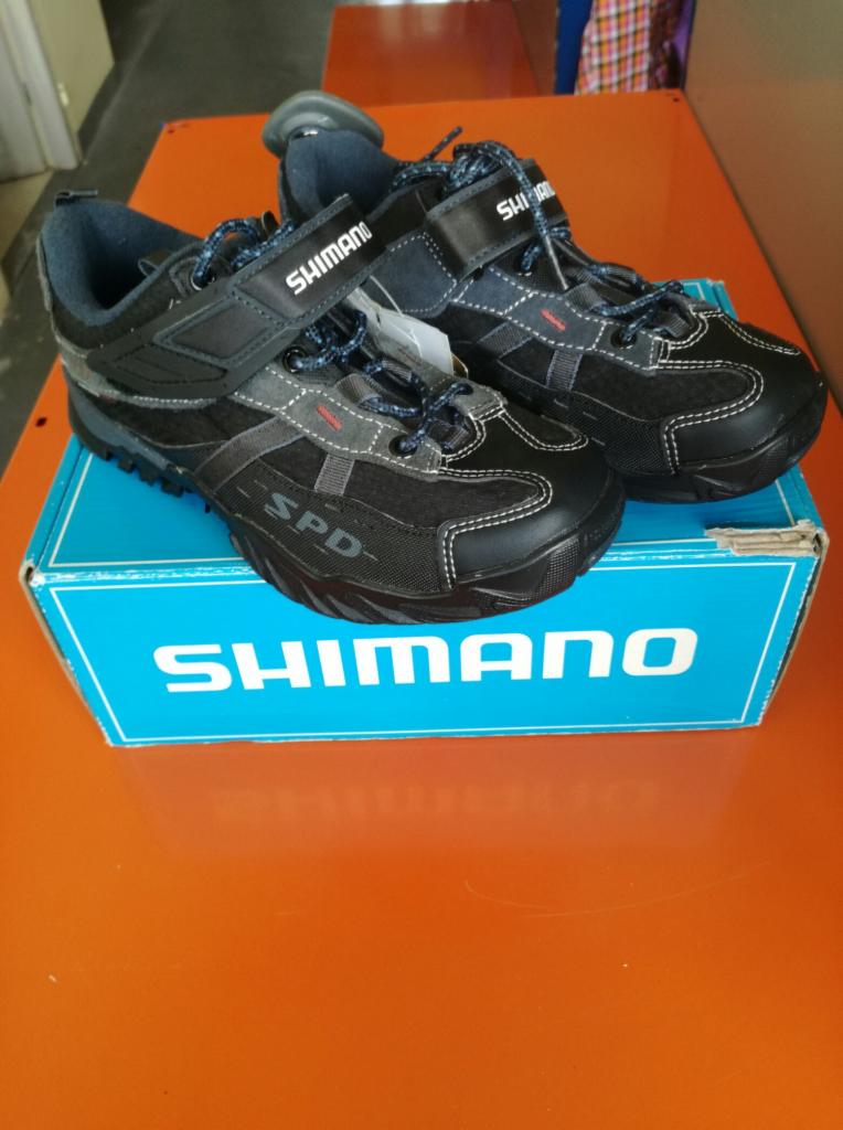 Para czarnych butów rowerowych Shimano SPD z niebieskimi sznurówkami, leżących na niebieskim pudełku z logo Shimano na pomarańczowym tle.