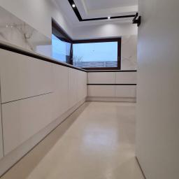 Usługi Budowlane Stanisław Bugajski - Minimalistyczna, nowoczesna kuchnia z białymi szafkami i blatem imitującym marmur, czarne akcenty na listwach i oknach, jasna podłoga żywiczna, widok na szary betonowy płot przez okno.