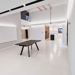 Usługi Budowlane Stanisław Bugajski - Przestronne, jasne wnętrze po remoncie, z białymi ścianami i podłogą, ciemnym stołem z marmurowym blatem na pierwszym planie, oraz widoczną kuchnią w tle z drewnianymi i białymi elementami.