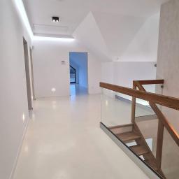 Usługi Budowlane Stanisław Bugajski - Jasne, przestronne wnętrze domu po remoncie, z białymi ścianami, podłogą z żywicy, drewnianymi schodami z szklaną balustradą i nowoczesnym oświetleniem sufitowym.