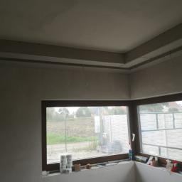 Usługi Budowlane Stanisław Bugajski - Wnętrze w trakcie prac wykończeniowych: sufit z zamontowaną konstrukcją z płyt kartonowo-gipsowych, widoczne przewody, ściany po nałożeniu gładzi, okno z widokiem na zewnątrz, na parapecie...