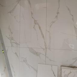 Usługi Budowlane Stanisław Bugajski - Fragment ściany wyłożonej białymi, błyszczącymi płytkami imitującymi marmur z widocznymi fugami, poziomica oparta o ścianę, oraz płytki przygotowane do montażu w prawym dolnym rogu.