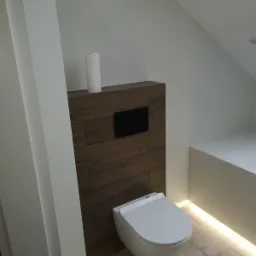 Nowoczesna toaleta podwieszana z deską wolnoopadającą zamontowana w łazience na poddaszu, obudowana płytami imitującymi drewno, białe płytki na ścianach i podświetlenie LED pod wanną.