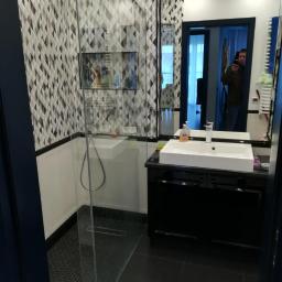 Usługi Budowlane Stanisław Bugajski - Nowoczesna łazienka z kabiną prysznicową walk-in, geometryczną mozaiką na ścianie, czarną szafką pod umywalką i dużym lustrem, w którym odbija się osoba robiąca zdjęcie.