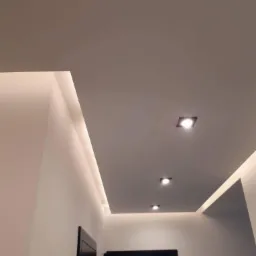 Nowoczesny sufit podwieszany z oświetleniem LED wzdłuż krawędzi i trzema punktowymi lampami, widok z dołu na tle białych ścian i czarnych drzwi.