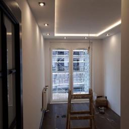 Usługi Budowlane Stanisław Bugajski - Wnętrze pokoju w trakcie remontu z drewnianą podłogą, białymi ścianami, sufitem podwieszanym z oświetleniem LED i drabiną stojącą przed oknem z widokiem na rusztowania na zewnątrz.