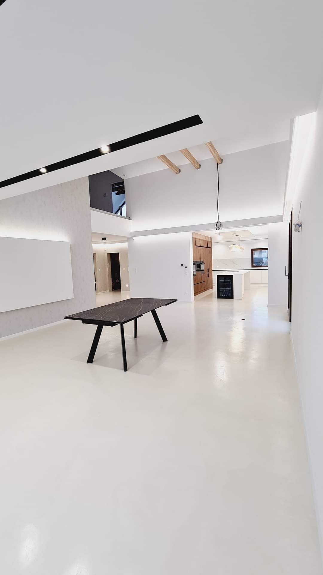 Przestronne, jasne wnętrze po remoncie, z białymi ścianami i podłogą, ciemnym stołem z marmurowym blatem na pierwszym planie, oraz widoczną kuchnią w tle z drewnianymi i białymi elementami.