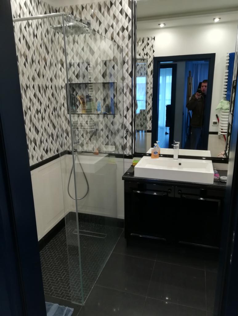Nowoczesna łazienka z kabiną prysznicową walk-in, geometryczną mozaiką na ścianie, czarną szafką pod umywalką i dużym lustrem, w którym odbija się osoba robiąca zdjęcie.
