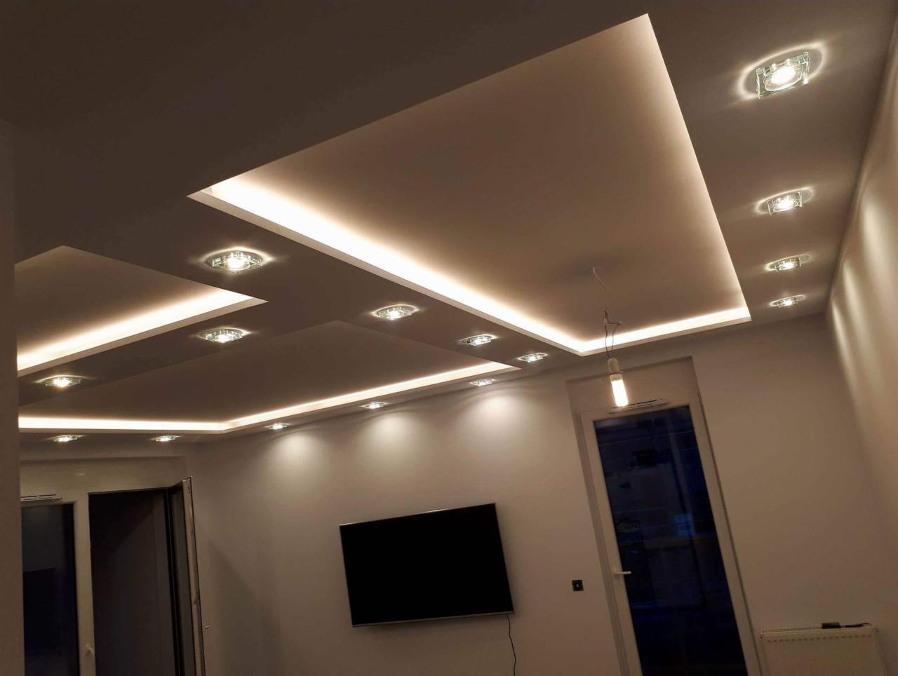 Nowoczesny salon z podwieszanym sufitem z oświetleniem LED i punktowymi lampami, widoczny telewizor na ścianie i minimalistyczna lampa wisząca.