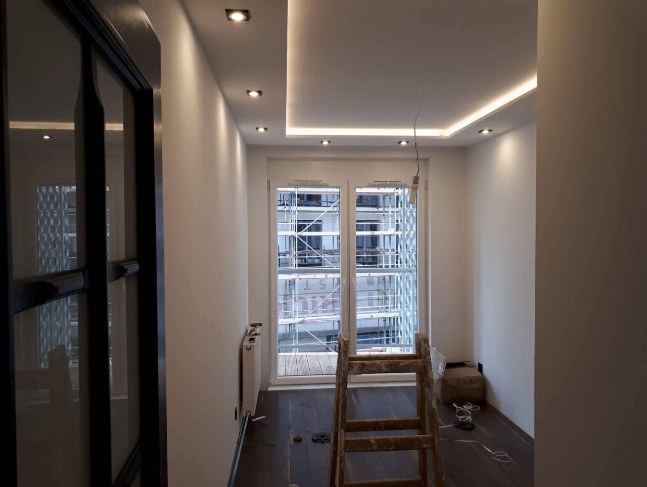 Wnętrze pokoju w trakcie remontu z drewnianą podłogą, białymi ścianami, sufitem podwieszanym z oświetleniem LED i drabiną stojącą przed oknem z widokiem na rusztowania na zewnątrz.