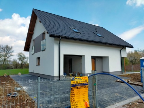 Nowoczesny dom z szarą dachówką i białymi ścianami, widoczne okna dachowe, balustrada balkonowa z szarego szkła, teren wokół domu częściowo wyłożony kostką, z przodu ogrodzenie budowlane i tablica...