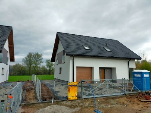 Nowoczesny, parterowy dom z ciemnym dachem i dwoma oknami dachowymi, ogrodzony metalową siatką, z widoczną przenośną toaletą budowlaną po prawej stronie, w tle zieleń i pochmurne niebo.