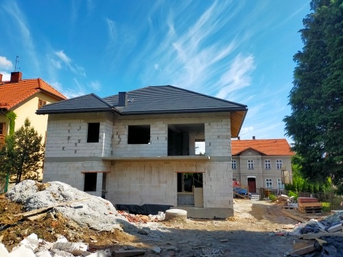 Surowy stan dwukondygnacyjnego domu z bloczków, z ciemną dachówką, na tle błękitnego nieba z chmurami i sąsiednich budynków z czerwonymi dachami.