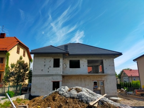 Dwukondygnacyjny dom w trakcie budowy, ściany z białych bloczków, czarny dach, widok na błękitne niebo z chmurami, sterta piasku przed budynkiem.