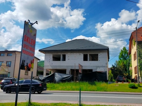 Surowy stan dwukondygnacyjnego domu z pustaków z ciemnoszarą dachówką, widoczny zza reklamy sklepu spożywczego przy ulicy.