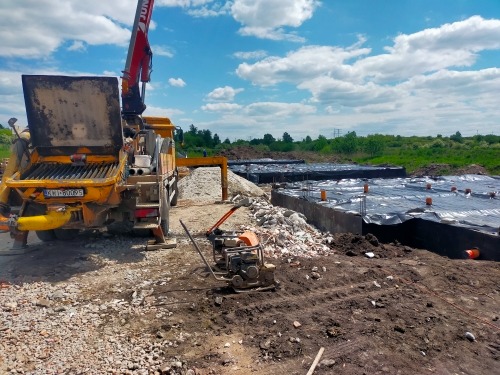 Fundamenty pod dom, pokryte folią, widoczna betoniarka z dźwigiem i ubijarka gruntu. Skotniki.